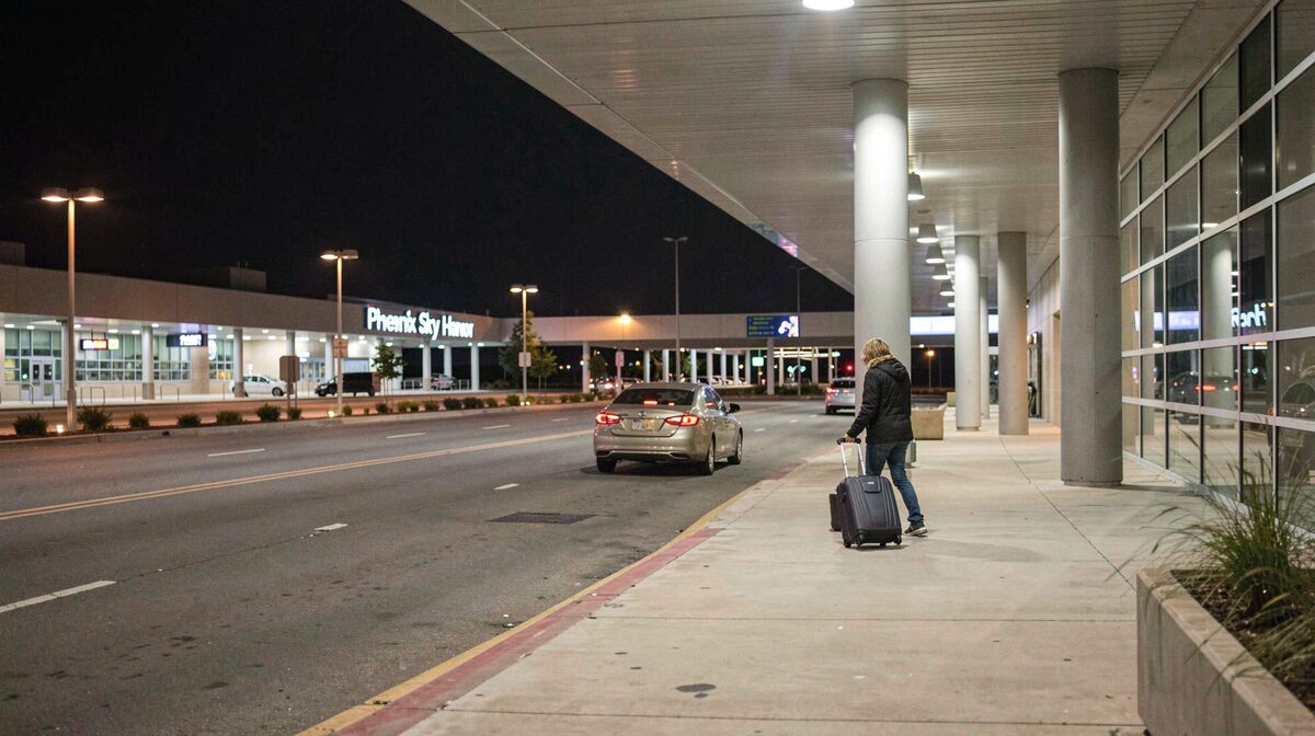 A nighttime view of a Phoenix Sky Harbor terminal curb lane, lit by overhead sodium lights, with a single rideshare car waiting and a traveler rolling a suitcase toward it, shot from the sidewalk at eye level.